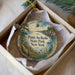A decorative ornament in a white box, featuring a scenic landscape with a body of water, trees, and a bridge.