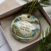 A decorative ornament in a box, featuring a scenic landscape with a body of water, mountains, and greenery.