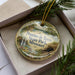 A decorative ornament in a box, featuring a scenic landscape with a waterfall, pine trees, and a river.