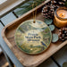 A decorative ornament featuring a scenic landscape of Prairie State Park, Missouri, with a bird perched on a tree branch. The ornament is placed on a wooden tray, surrounded by pine cones and a lit candle, creating a cozy and festive atmosphere.