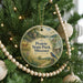 A round ornament hanging on a Christmas tree, featuring a scenic landscape with a bird, a cabin, and a river.