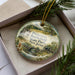 A decorative ornament in a box, featuring a scenic landscape with a house, bridge, and trees.