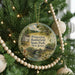 A round ornament hanging on a Christmas tree, featuring a scenic landscape with a house, bridge, and trees.