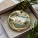 A decorative ornament in a box, featuring a scenic landscape with mountains, trees, and a bird in flight.