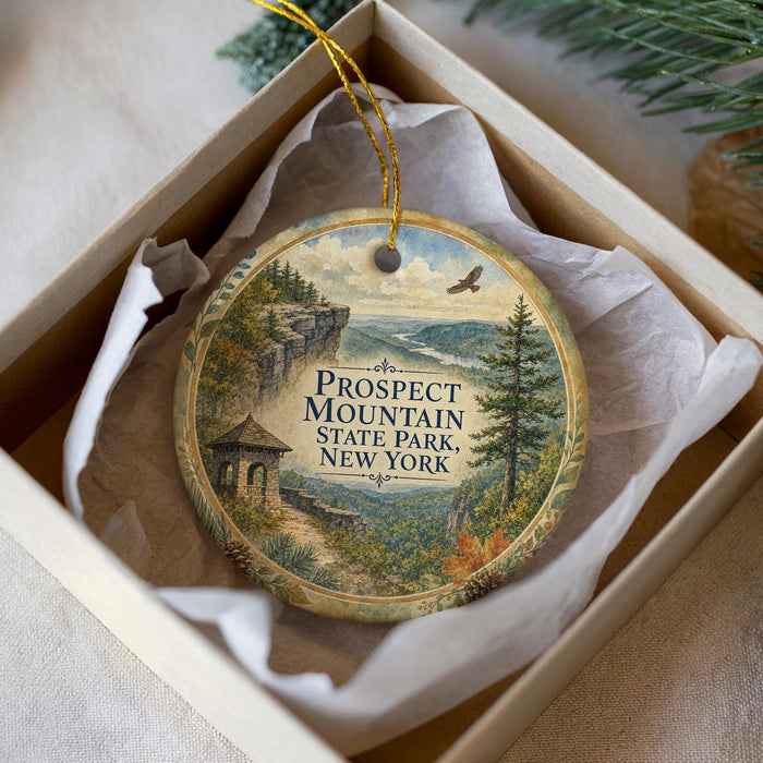 A decorative ornament in a box, featuring a scenic landscape with a mountain, a cabin, and a bird in flight.