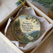 A decorative ornament in a white box, featuring a scenic landscape with mountains, pine trees, and a river.