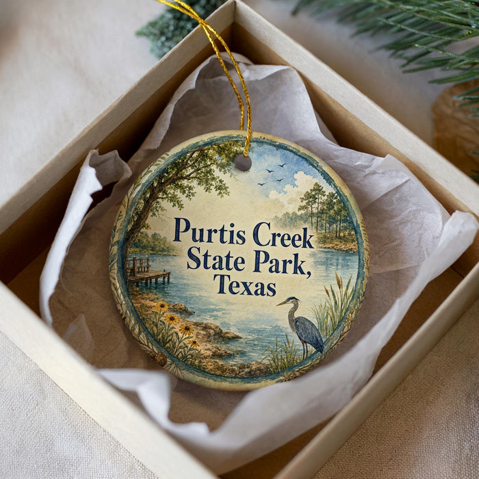 A decorative ornament in a white box, featuring a painting of a bird standing on a rock near a body of water, with the text "Purtis Creek State Park, Texas" displayed on the ornament.