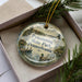 A decorative ornament in a box, featuring a scenic image of a lake and a lighthouse, with the text "Pymatuning State Park, Pennsylvania" printed on it.