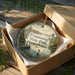 A decorative ornament in a box, featuring a scenic landscape with a lake, trees, and ducks, along with the text "Pymatuning State Park, Pennsylvania".