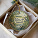 A round ornament with a painting of a lake and a bird, placed in a box with a white tissue paper lining.