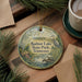 A decorative ornament with a bird and a lake scene, surrounded by pine branches and a cup of coffee on a wooden surface.