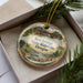 A decorative ornament in a box, featuring a scenic landscape with a bridge and pine trees.