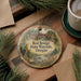 A decorative ornament featuring a scenic landscape with a bridge, surrounded by pine branches and flowers, placed on a wooden surface alongside a cup of coffee.