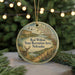 A round ornament with a scenic landscape of a river, mountains, and wildlife, including a deer, is displayed in front of a blurred background of Christmas lights.
