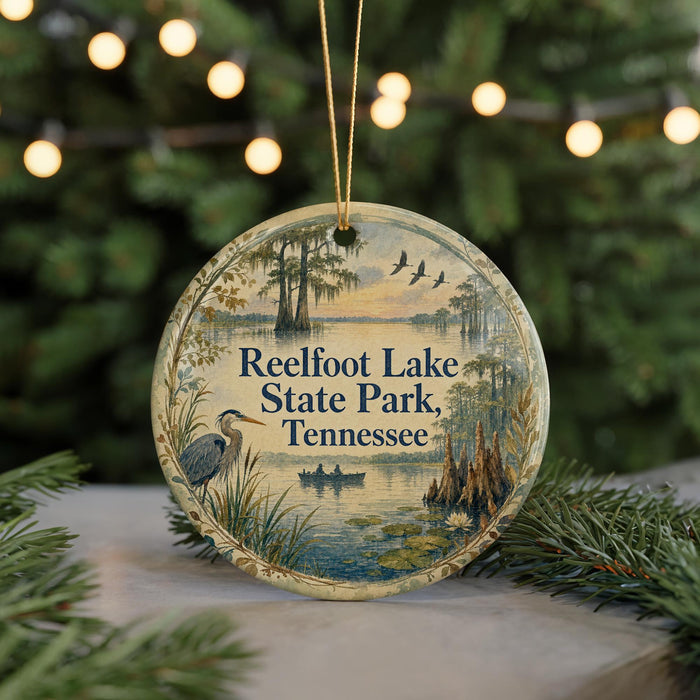 A circular ornament with a painting of a lake and birds, surrounded by greenery, hangs in front of a Christmas tree with lights.
