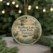 A circular ornament with a painting of a lake and birds, surrounded by greenery, hangs in front of a Christmas tree with lights.