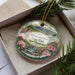 A decorative ornament with a floral design and text, placed in a box with pine branches.