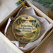 A round ornament with a scenic landscape and text, placed in a box with a white tissue paper lining.