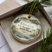 A decorative ornament in a box, featuring a scenic image of a lake surrounded by mountains and pine trees.