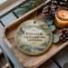 A wooden tray with a decorative ornament featuring a scenic lake and pine trees, along with a lit candle and pine cones.