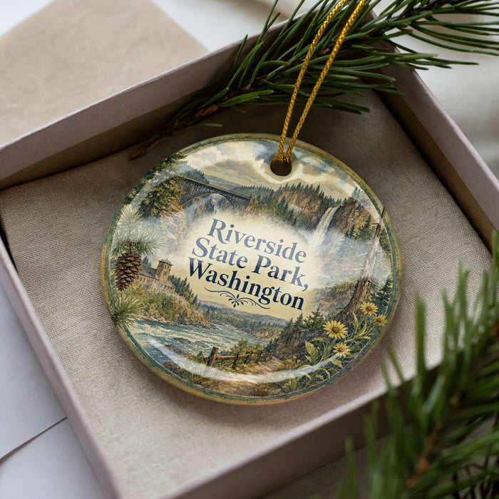 A decorative ornament in a box, featuring a scenic landscape with a river, mountains, and pine trees.