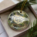 A decorative ornament in a box, featuring a scenic landscape with a waterfall and a bridge.