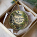 A round ornament with a scenic image of a waterfall and a bridge, placed in a box with a white tissue paper lining.
