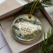 A decorative ornament in a box, featuring a scenic landscape with a river, bridge, and people enjoying outdoor activities.