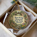 A round ornament with a scenic illustration of a bridge, trees, and a bird, placed in a white box with a gold string.