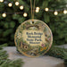 A round ornament with a scenic landscape of a river, cabin, and trees, featuring a bird perched on a branch, is displayed in front of a blurred background with twinkling lights.