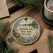 A round ornament with a scenic lake and park landscape, featuring a bird, is displayed on a wooden surface alongside a cup of coffee and some pine branches.