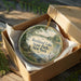 A round ornament in a wooden box, featuring a scenic landscape with a lake and trees, with the text "Rockland Lake State Park, New York" printed on it.