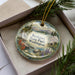 A decorative ornament in a box, featuring a scenic landscape with mountains, water, and pine trees.