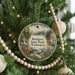 A round ornament hanging on a Christmas tree, featuring a scenic landscape with mountains, a waterfall, and a cabin.