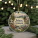 A round ornament with a scenic landscape of a lake, mountains, and a cabin, surrounded by pine trees and colorful flowers, hangs in front of a blurred Christmas tree with lights.
