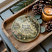 A wooden tray with a decorative ornament featuring a house and a tree, surrounded by pine cones and a lit candle.