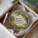 A round ornament with a vintage-style illustration of a house and a tree, placed in a white box with a gold string.
