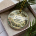 A decorative ornament in a box, featuring a vintage-style illustration of a house surrounded by trees and a fence, with the text "Rose Hill Plantation State Historic Site, South Carolina" displayed on it.