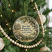 A round ornament hanging on a Christmas tree, featuring a scenic image of a house surrounded by trees and a pond.