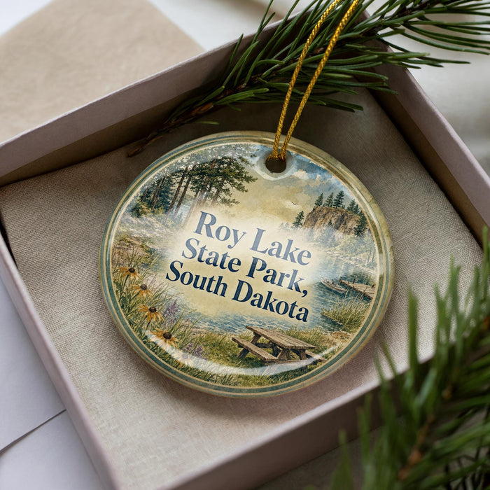 A decorative ornament in a box, featuring a scenic landscape with pine trees, a lake, and a picnic table.