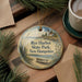 A decorative ornament featuring a lighthouse and a boat, with the text "Rye Harbor State Park, New Hampshire" displayed on it. The ornament is placed on a wooden surface, surrounded by pine branches and a cup of coffee.
