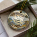 A decorative ornament with a landscape scene, featuring mountains, trees, and a body of water, is displayed in a box with pine branches.
