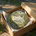 A decorative ornament in a box, featuring a scenic landscape with a house, trees, and a waterfall.
