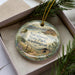 A decorative ornament in a box, featuring a scenic landscape with mountains, trees, and a cabin.