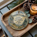 A decorative ornament featuring a scenic mountain landscape with pine trees and a bird perched on a branch, placed on a wooden tray alongside pine cones and a lit candle.