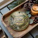 A round ornament with a scenic image of Salt Springs State Park, Pennsylvania, featuring a waterfall and a house, is displayed on a wooden tray alongside pine cones and a lit candle.