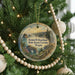 A round ornament hanging on a Christmas tree, featuring a scenic landscape with mountains, a river, and a bridge.