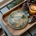 A decorative ornament with a scenic landscape of Sand Bar State Park, Vermont, featuring a lake, mountains, pine trees, and purple flowers. The ornament is displayed on a wooden tray, accompanied by pine cones and a lit candle.