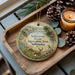 A decorative ornament featuring a scenic landscape of the state of Nebraska, with a deer and pine cones on a wooden tray.