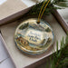 A decorative ornament in a box, featuring a scenic landscape with mountains, pine trees, and a cabin.